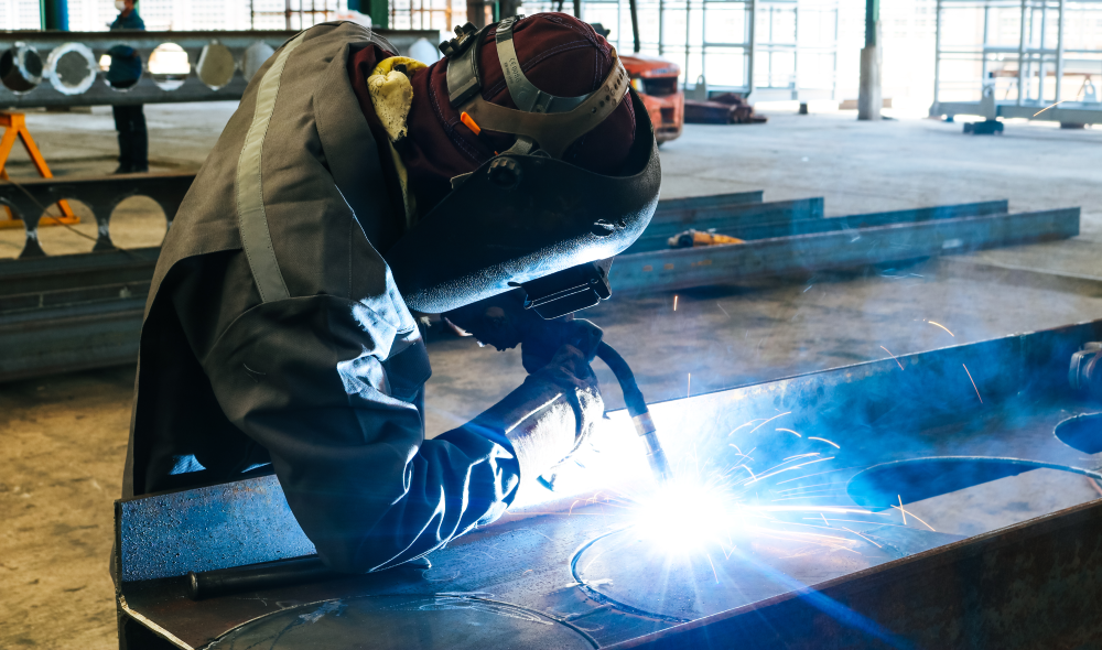 A welder wearing protective gear works on metal, creating bright sparks and a blue arc, in a spacious industrial workshop